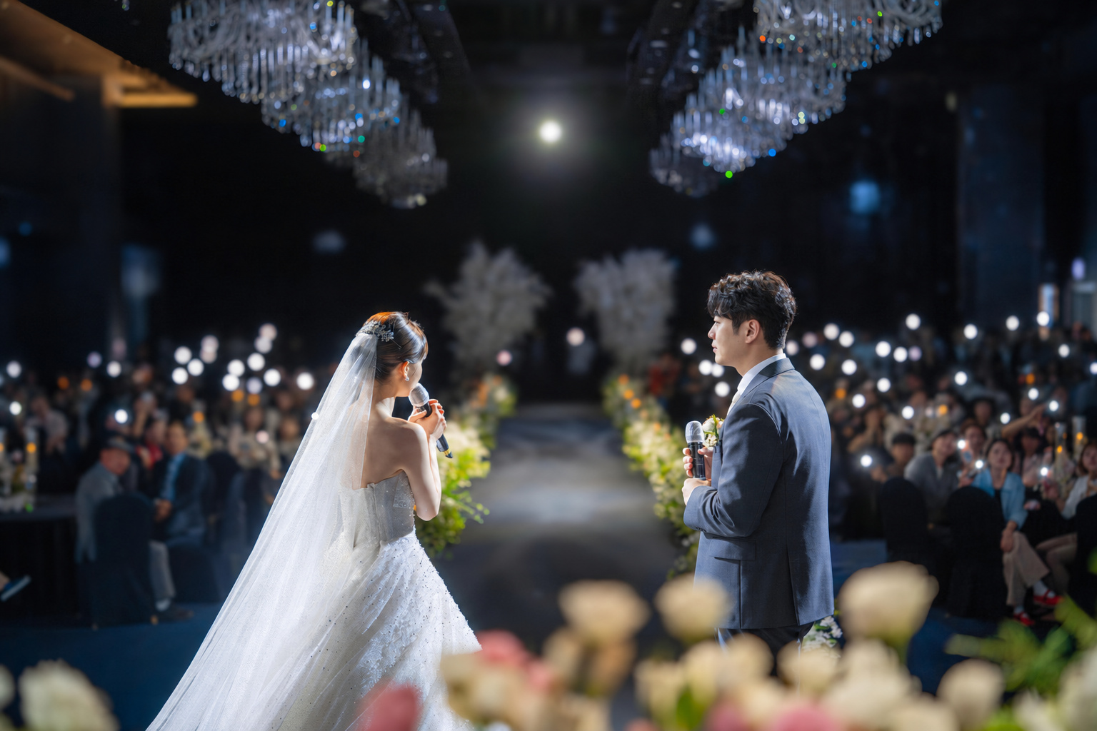 하객들의 불빛 속에서 마이크를 들고 서로를 바라보는 신랑과 신부